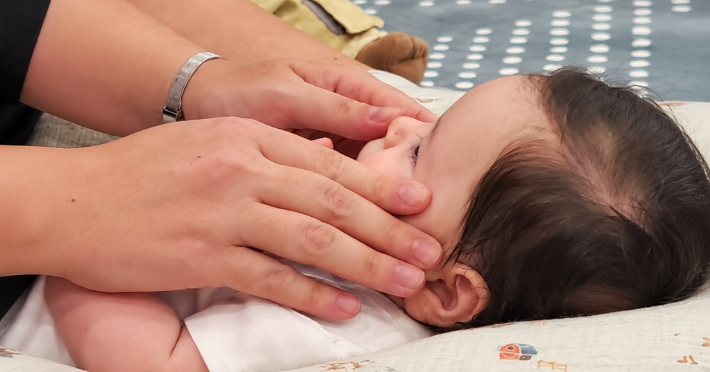 Mani adulte accarezzano il viso di un bebè durante un momento di massaggio infantile.